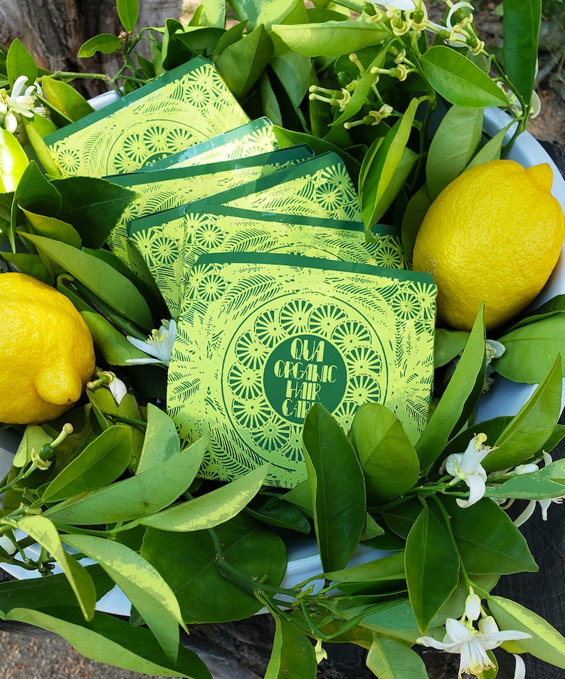 Puede incluir: Una pila de tarjetas con patrones verdes y amarillos con el texto "QUA ORGANIC HAIR CARE" en la tarjeta superior. Las tarjetas est&aacute;n rodeadas de hojas verdes y flores blancas.