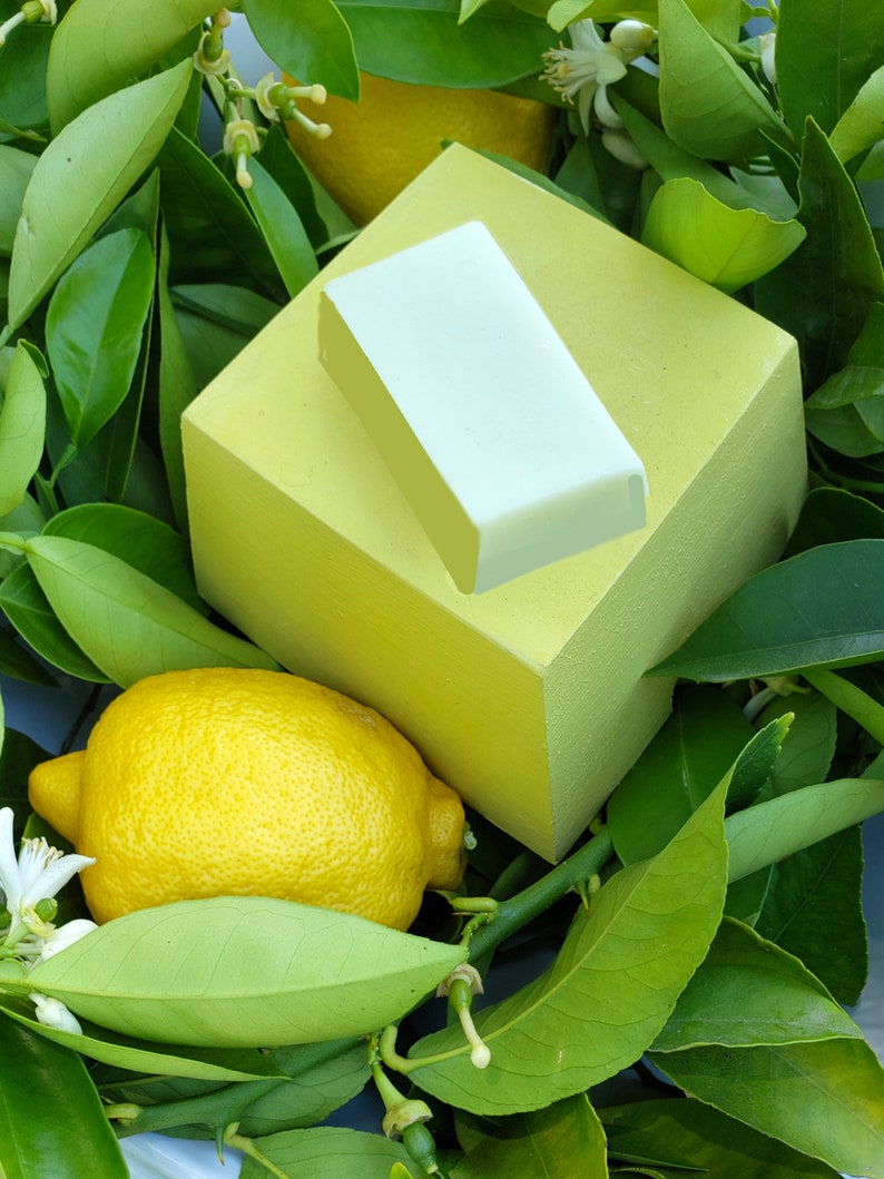Puede incluir: Un lim&oacute;n amarillo se encuentra sobre un lecho de hojas verdes, junto a una pastilla de jab&oacute;n cuadrada de color verde claro. La pastilla de jab&oacute;n est&aacute; sobre un cuadrado amarillo m&aacute;s grande.