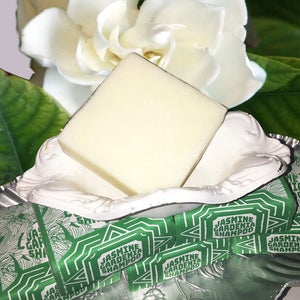 Puede incluir: Una barra de jab&oacute;n blanca en un plato blanco y plateado con un embalaje verde y blanco que dice "Champ&uacute; de Jazm&iacute;n y Gardenia". El plato est&aacute; sobre una superficie blanca con una flor blanca detr&aacute;s.