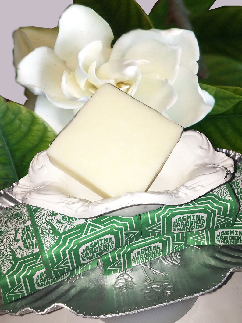 Puede incluir: Una barra de jab&oacute;n blanca en un plato blanco y plateado con un embalaje verde y blanco que dice "Champ&uacute; de Jazm&iacute;n y Gardenia". El plato est&aacute; sobre una superficie blanca con una flor blanca detr&aacute;s.