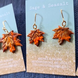 May include: A pair of gold dangle earrings with orange and brown maple leaf charms. The earrings are handmade in England and are available for purchase on Etsy.