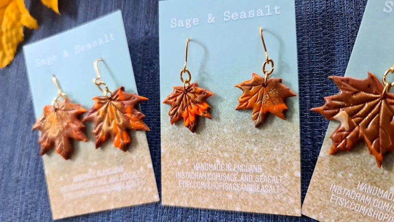 May include: A pair of gold dangle earrings with orange and brown maple leaf charms. The earrings are handmade in England and are available for purchase on Etsy.