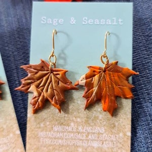 May include: A pair of gold dangle earrings with orange and brown maple leaf charms. The earrings are handmade in England and are available on Etsy and Instagram. The text "Sage & Seasalt" is printed on the card behind the earrings.