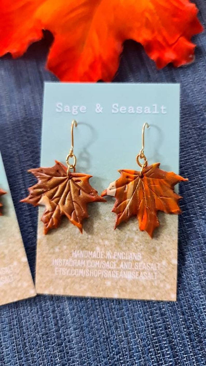 May include: A pair of gold dangle earrings with orange and brown maple leaf charms. The earrings are handmade in England and are available on Etsy and Instagram. The text "Sage & Seasalt" is printed on the card behind the earrings.