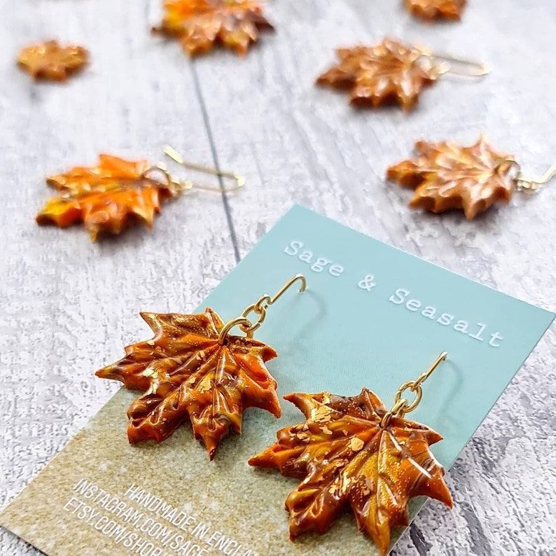 May include: A pair of gold dangle earrings with orange and gold maple leaf charms. The earrings are handmade in England and are available on Etsy and Instagram. The card reads "Sage & Seasalt".