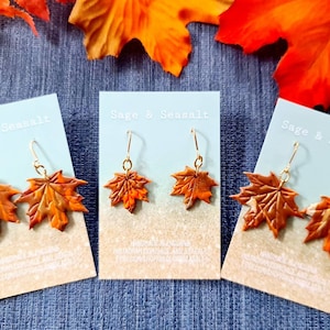 May include: Three pairs of brown maple leaf earrings on white cards with the text "Sage & Seasalt" printed on them. The cards are on a blue fabric background.