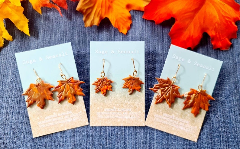 May include: Three pairs of brown maple leaf earrings on white cards with the text "Sage & Seasalt" printed on them. The cards are on a blue fabric background.