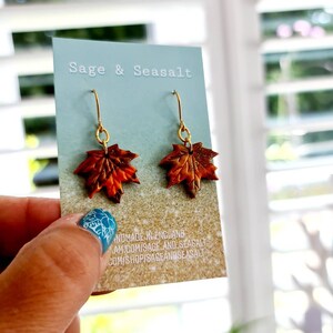 May include: A pair of gold dangle earrings with autumn-colored maple leaf charms. The leaves are a mix of orange, brown, and red with a glittery finish. The card reads "Sage & Seasalt Handmade in England sageandseasalt.com/shop/sageandseasalt"