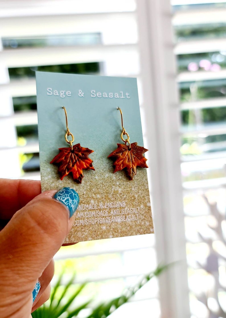 May include: A pair of gold dangle earrings with autumn-colored maple leaf charms. The leaves are a mix of orange, brown, and red with a glittery finish. The card reads "Sage & Seasalt Handmade in England sageandseasalt.com/shop/sageandseasalt"