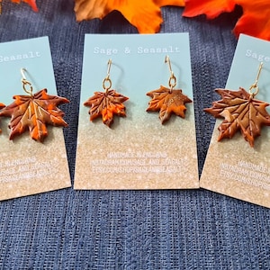 May include: Three pairs of dangle earrings featuring maple leaf designs. The leaves are made of polymer clay and are painted in shades of orange, brown, and gold. The earrings are displayed on cards with the text "Sage & Seasalt" and information about the shop's social media accounts.