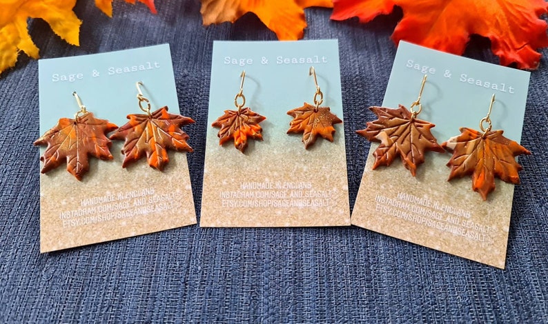 May include: Three pairs of dangle earrings featuring maple leaf designs. The leaves are made of polymer clay and are painted in shades of orange, brown, and gold. The earrings are displayed on cards with the text "Sage & Seasalt" and information about the shop's social media accounts.