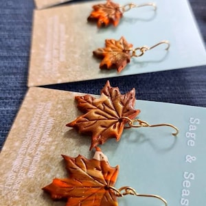 May include: A pair of gold dangle earrings featuring autumn-colored maple leaf charms. The leaves are made of polymer clay and have a textured, painted finish. The earrings are handmade and come from Sage & Seasalt.