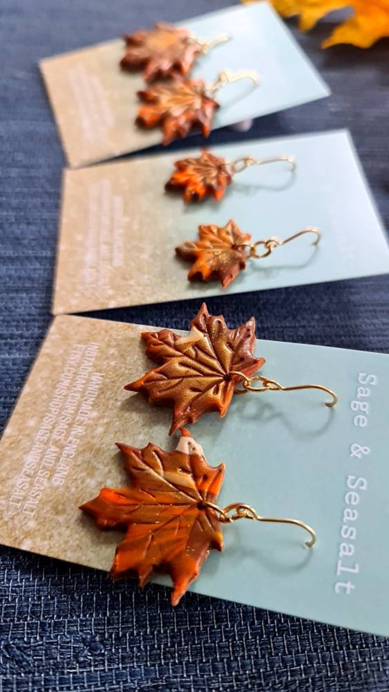 May include: A pair of gold dangle earrings featuring autumn-colored maple leaf charms. The leaves are made of polymer clay and have a textured, painted finish. The earrings are handmade and come from Sage & Seasalt.