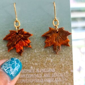 May include: A pair of gold dangle earrings with orange, brown, and gold maple leaf charms. The earrings are displayed on a card with the text "Sage & Seasalt" and "Handmade in England".