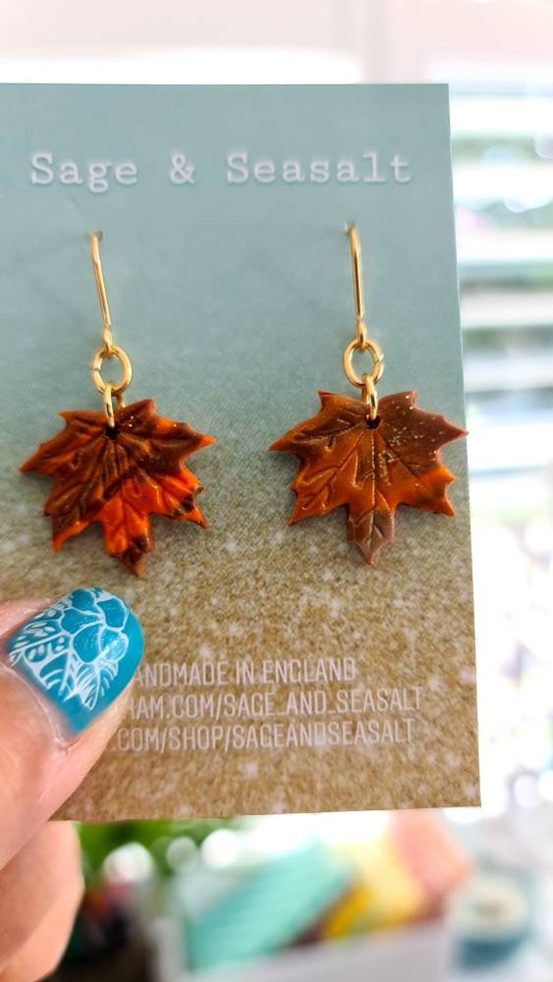May include: A pair of gold dangle earrings with orange, brown, and gold maple leaf charms. The earrings are displayed on a card with the text "Sage & Seasalt" and "Handmade in England".