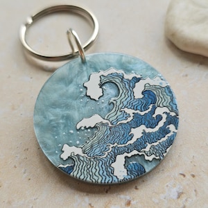 May include: A circular keyring with a silver ring. The keyring showcases a blue and white wave design set against a light blue backdrop. The wave design is detailed, with white foam and dark blue lines. This keyring is a decorative accessory.