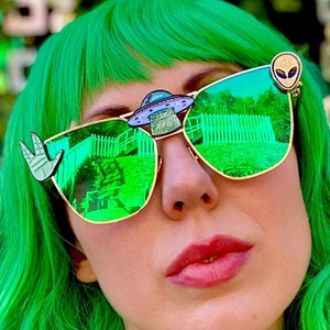 Op de afbeelding: Een vrouw met felgroen haar draagt een groene, gespiegelde zonnebril met buitenaardse accenten. Ze draagt ook een zilveren choker met de letters "A&B" erop.