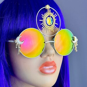 May include: A pair of round, iridescent rainbow sunglasses with gold metal frames and dangling crescent moon charms. The sunglasses are worn by a person with bright purple hair and a gold sun and star headpiece.
