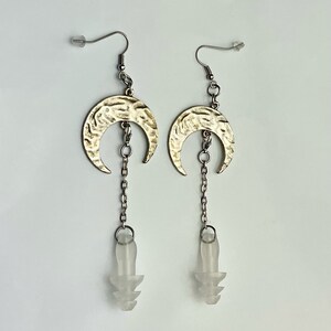 May include: A pair of silver-toned earrings featuring a crescent moon design with a hammered texture. Each earring has a chain that connects to a clear, tapered plug. The earrings have a hook closure.