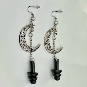 May include: A pair of silver-toned crescent moon earrings with intricate filigree detailing. Each earring features a dangling black, layered plug design, suspended by a delicate chain. The earrings have a hook closure.
