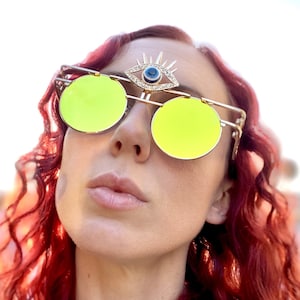May include: A woman with red hair wearing round, yellow-tinted sunglasses with a gold frame. The sunglasses have a decorative third eye with a blue stone and rhinestones on the bridge.