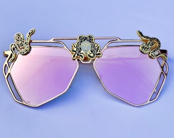 Peut inclure: Lunettes de soleil dorées avec des verres miroirs roses et des accents décoratifs de serpent et de Méduse.