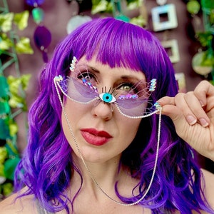 May include: A woman with bright purple hair is wearing a pair of iridescent butterfly-shaped sunglasses with a blue eye charm. The sunglasses have a silver chain attached to them.