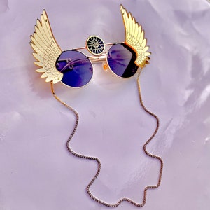 May include: Gold-tone sunglasses with a chain, featuring a winged design on each side of the frame. The lenses are dark and round. The bridge of the glasses has a small, round, gold-tone charm with a black eye symbol.