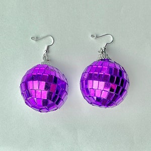 May include: A pair of purple disco ball earrings with silver hooks. The earrings are made of small mirrored tiles.