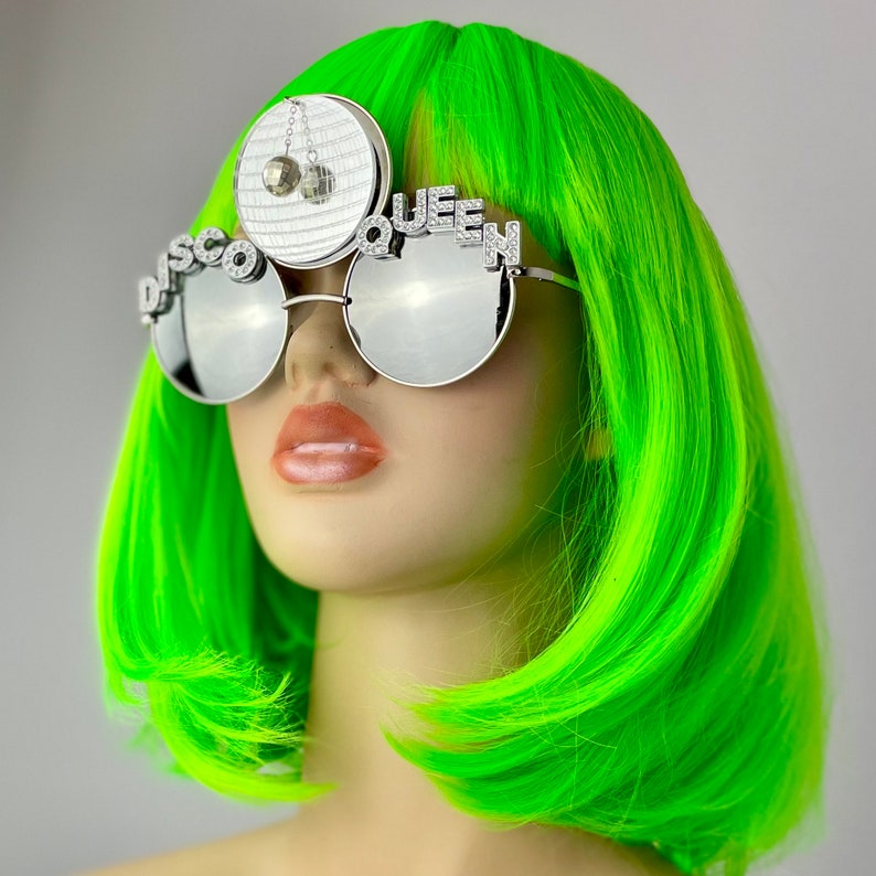 Może przedstawiać: Srebrne okulary przeciwsłoneczne z lustrzanymi szkłami z napisem "Disco Queen" na oprawce. Okulary nosi manekin w jasnozielonej peruce.