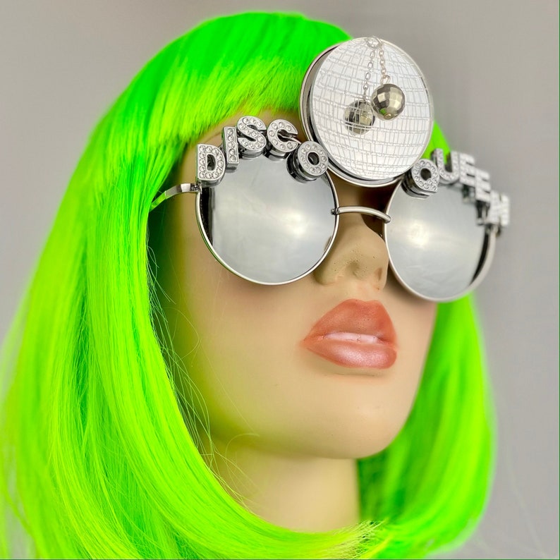 Może przedstawiać: Srebrne okulary przeciwsłoneczne z lustrzanymi szkłami, z napisem "Disco Queen" wysadzanym kryształami. Okulary nosi manekin z jasnozielonymi włosami i srebrną dyskoteką na głowie.