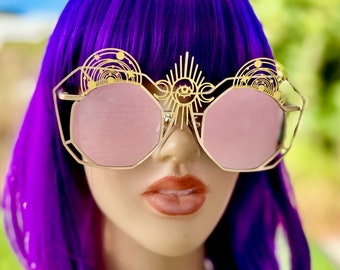 Peut inclure: Lunettes de soleil dorées avec des verres roses et un motif céleste. Les lunettes de soleil présentent un motif de soleil éclatant avec un œil au centre et un motif céleste sur les branches.