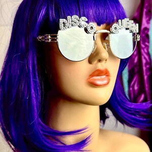 Może przedstawiać: Srebrne okulary przeciwsłoneczne z napisem "Disco Queen" wysadzanym cyrkoniami. Okulary mają duże, okrągłe szkła i są noszone przez manekina z fioletowymi włosami.