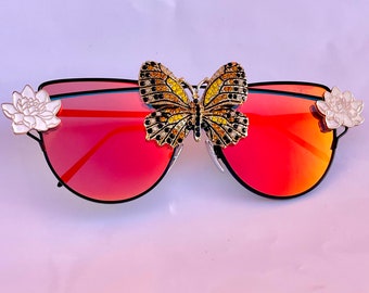 Peut inclure: Lunettes de soleil de forme œil de chat à miroir rouge avec un accent papillon en strass et des embellissements de fleurs blanches sur les côtés.