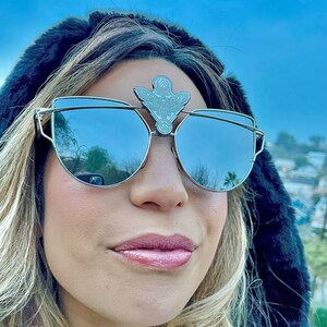 Könnte beinhalten: Sonnenbrille mit silbernem Rahmen, verspiegelten Gläsern und einem dekorativen silbernen Emblem über dem Steg. Die Sonnenbrille hat ein einzigartiges, eckiges Design. Die Gläser spiegeln den Himmel und die Umgebung wider. Der Gesamtstil ist modern und modisch.