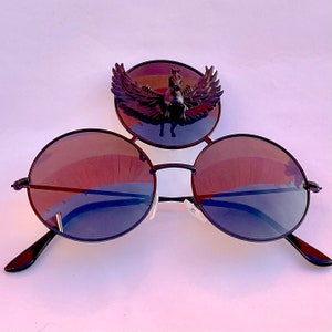 May include: Black round sunglasses with a decorative black phoenix design on the bridge. The sunglasses have a dark brown lens.