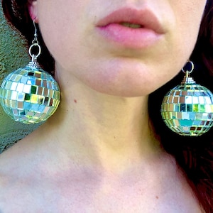 May include: A pair of silver disco ball earrings with a mirrored surface. The earrings are large and round, and they are hanging from the ears of a person.