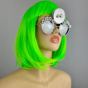 Może przedstawiać: Srebrne okulary przeciwsłoneczne "Disco Queen" z lustrzanymi szkłami i zawieszką w kształcie kuli dyskotekowej. Okulary mają napis "Disco Queen" na oprawce.
