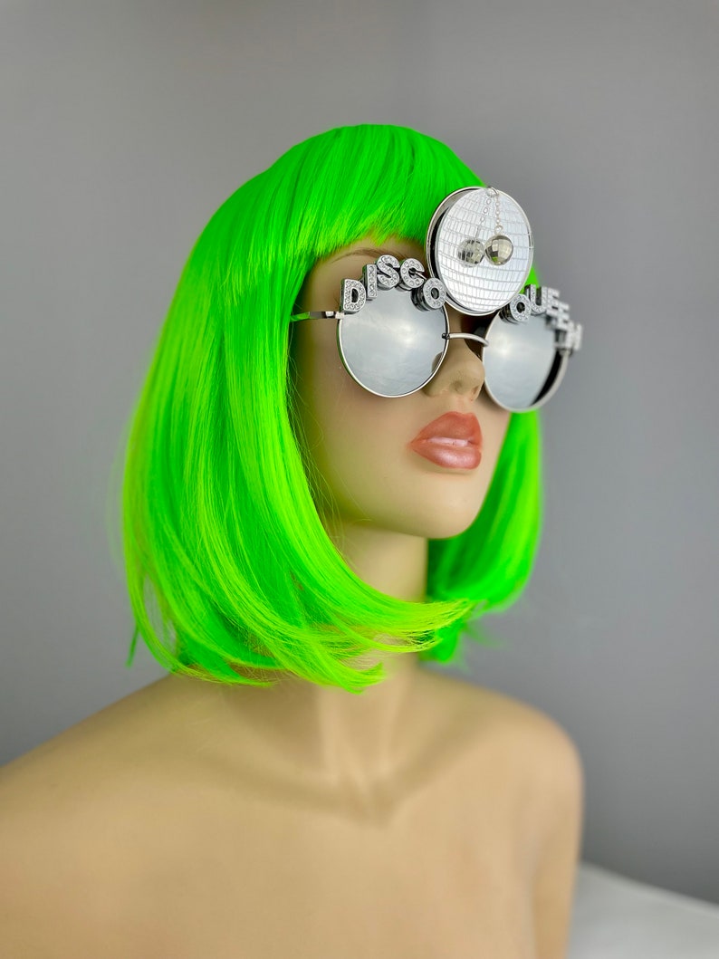 Może przedstawiać: Srebrne okulary przeciwsłoneczne "Disco Queen" z lustrzanymi szkłami i zawieszką w kształcie kuli dyskotekowej. Okulary mają napis "Disco Queen" na oprawce.