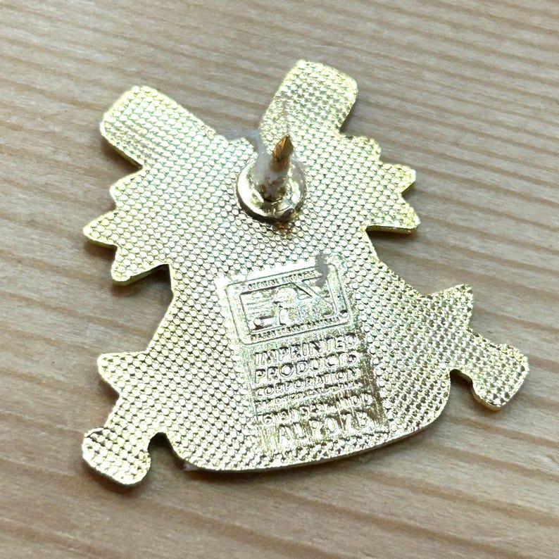 May include: A gold-colored pin with a textured surface and a pin backing. The pin has a unique shape with multiple points and a rectangular stamp with text. The pin is on a light brown wooden surface.