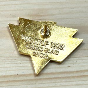 May include: A gold-toned metal lapel pin with a triangular, arrow-like shape. The back features a central pin post and is embossed with the text TM/&copy; NFLP 1992 GRAND SLAM CHINA. The surface has a textured, metallic finish with visible stamped lettering.