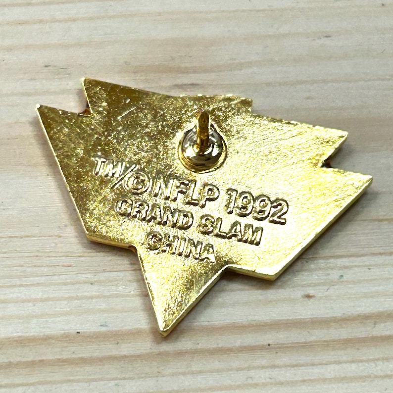 May include: A gold-toned metal lapel pin with a triangular, arrow-like shape. The back features a central pin post and is embossed with the text TM/&copy; NFLP 1992 GRAND SLAM CHINA. The surface has a textured, metallic finish with visible stamped lettering.