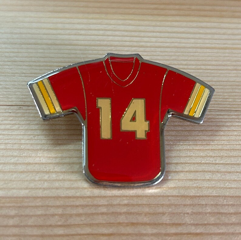 Puede incluir: Un pin de camiseta de f&uacute;tbol roja y dorada con el n&uacute;mero 14 en la parte delantera.