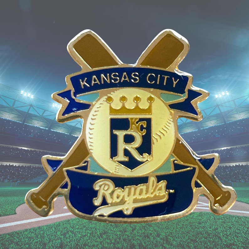 May include: A gold and blue Kansas City Royals baseball pin. The pin features the team's logo, a baseball with a crown, the letter "R", and crossed bats. The words "Kansas City" and "Royals" are displayed on blue banners.