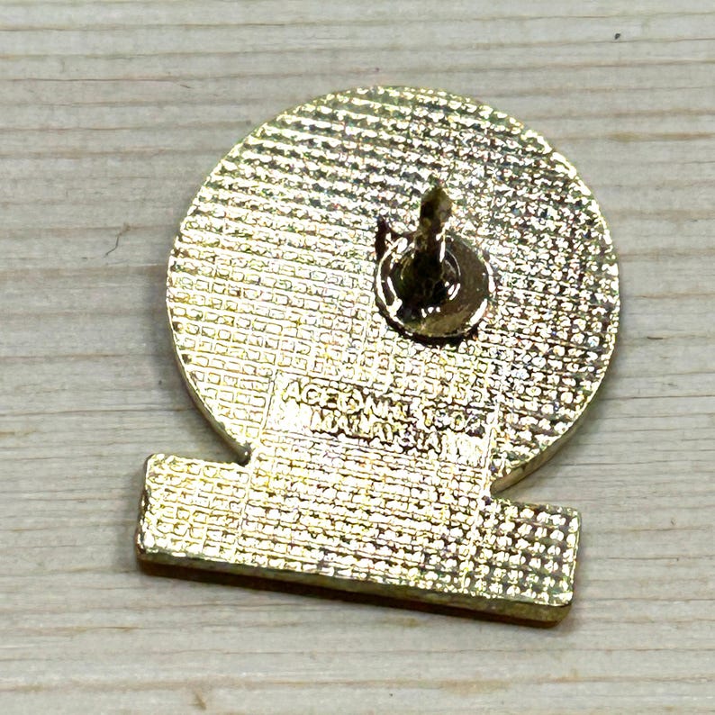 Puede incluir: Un pin dorado con una parte superior redonda y una inferior rectangular. El pin tiene una superficie texturizada y un cierre de alfiler. El texto "ACE SDN BHD MALAYSIA" es visible en el pin.