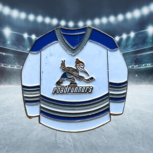 May include: A hockey jersey-shaped pin with a white and blue design. The pin features a cartoon roadrunner playing hockey, with the word "roadrunners" below. The jersey has blue and gray accents.