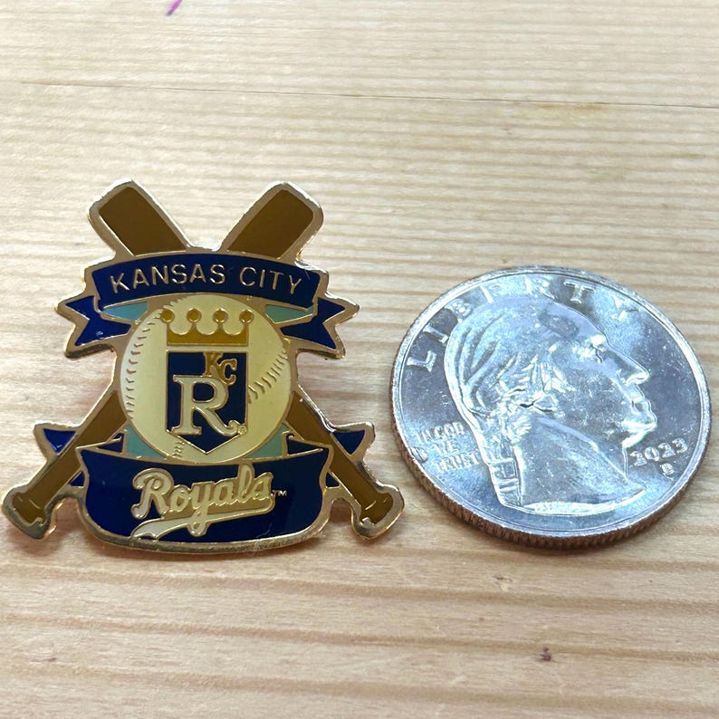 May include: A Kansas City Royals baseball team pin with a baseball and crossed bats design. The pin is gold, blue, and white. A US quarter coin is next to the pin.