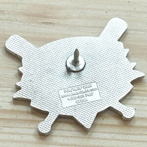 May include: A silver-colored pin with a baseball-themed design, featuring a baseball and crossed bats. The pin has a textured surface and a pin backing. The text on the pin reads "POL/MEP 2006" and includes a website and phone number.