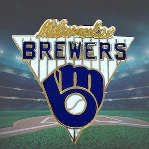 May include: A triangular enamel pin featuring the text Milwaukee Brewers in gold and blue lettering over a white pinstriped background. Below the text is a large blue baseball glove logo with a white baseball centered in the palm area.