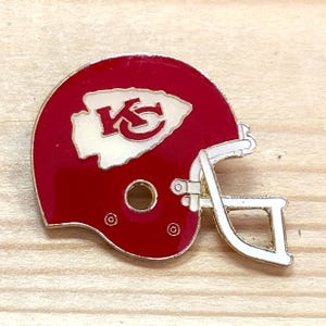 May include: A red and white enamel pin in the shape of a football helmet. The helmet features the team's logo in white on the side. The pin has a gold-colored outline and a pin back.